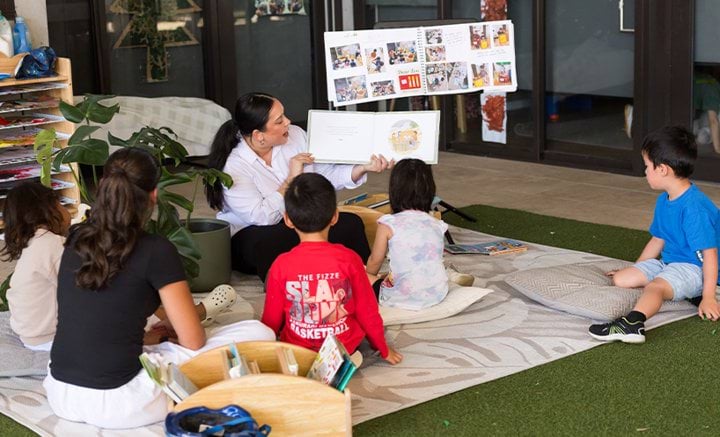 Two educators sitting on the floor reading a book to a group of children.