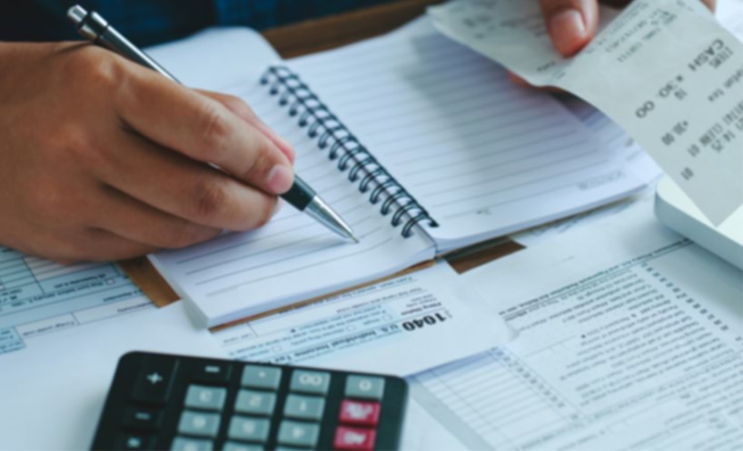 A person at a desk writing in a notepad with a calculator and paperwork work.